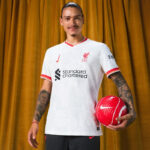 Smiling man in a white Liverpool FC jersey holding a red Nike football against a gold curtain backdrop.