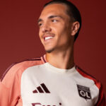 Portrait of a smiling man in a white-pink soccer jersey with the OL Lyonnais crest against a red background.