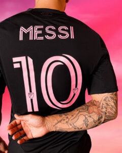 Back view of a black jersey with pink 'MESSI' text and large '10', MLS logos, worn by a tattooed person against a pink gradient background.