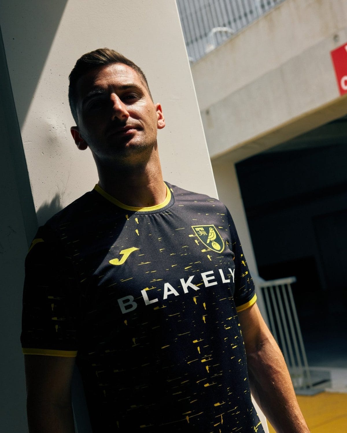 Man leaning against a light wall outdoors, wearing a black and yellow sports jersey with 'BLAKELY' text and crest on the chest.