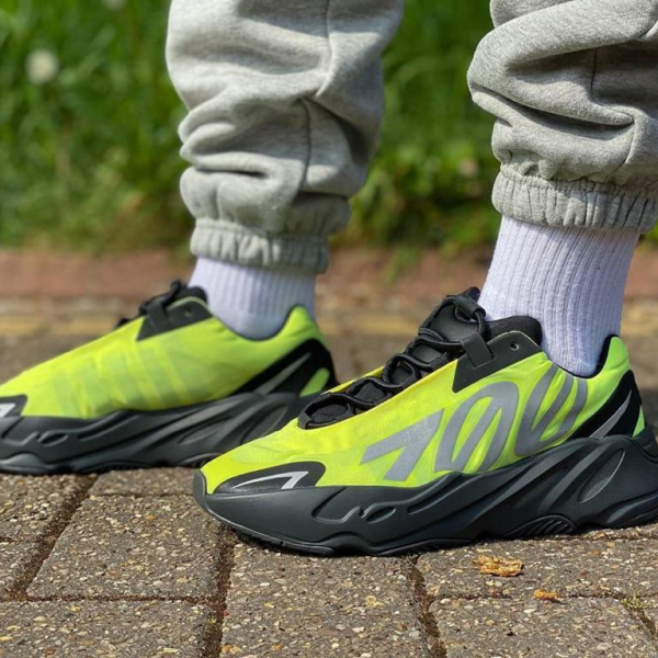 Neon yellow-green sneakers with a chunky black sole on a person standing on brick pavement.