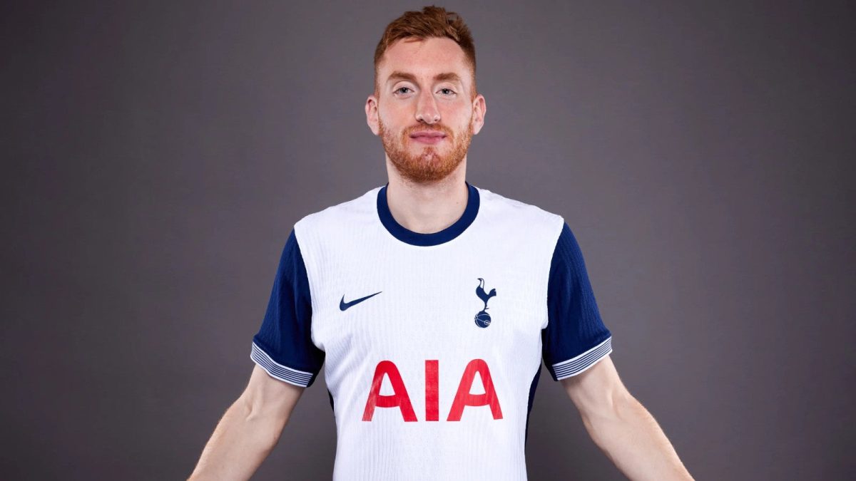 Man with short red hair and beard wearing a white Tottenham jersey with blue sleeves and red 'AIA' sponsor logo, standing against a gray backdrop.