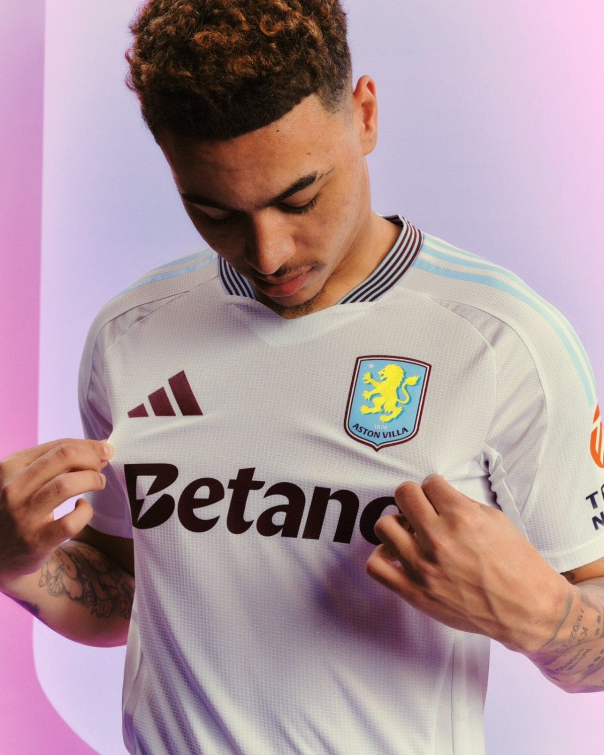 Man in an Aston Villa football jersey looks down while tugging at the shirt, against a pink-purple background.