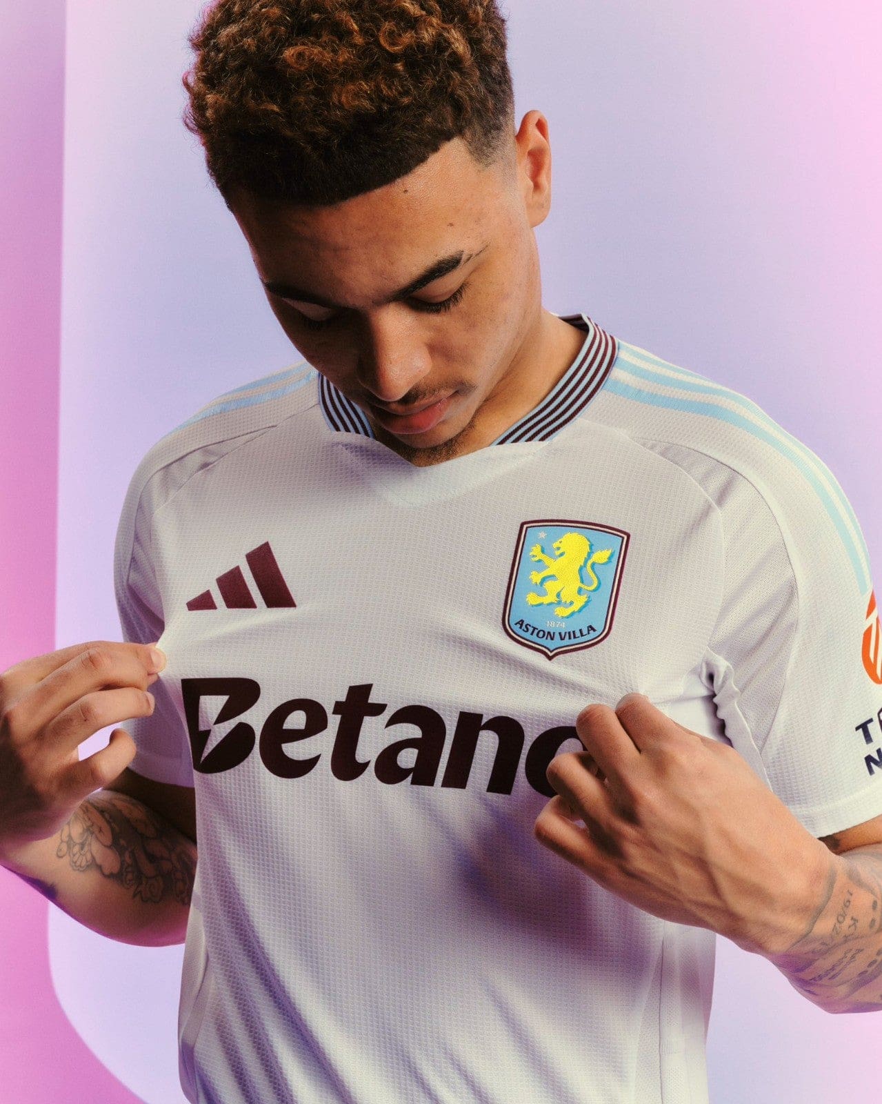 i6lAhTgq8d0N2AC | UAE SPORT STORE Man in an Aston Villa football jersey looks down while tugging at the shirt, against a pink-purple background.