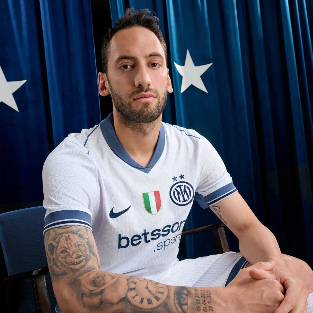 Portrait of a male soccer player in a white Inter Milan jersey with crest and Italian flag badge, seated with tattooed forearms visible against a blue curtain backdrop.