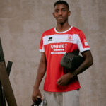 Young man in red and white soccer jersey holding a black crossbody bag, standing against a neutral backdrop.