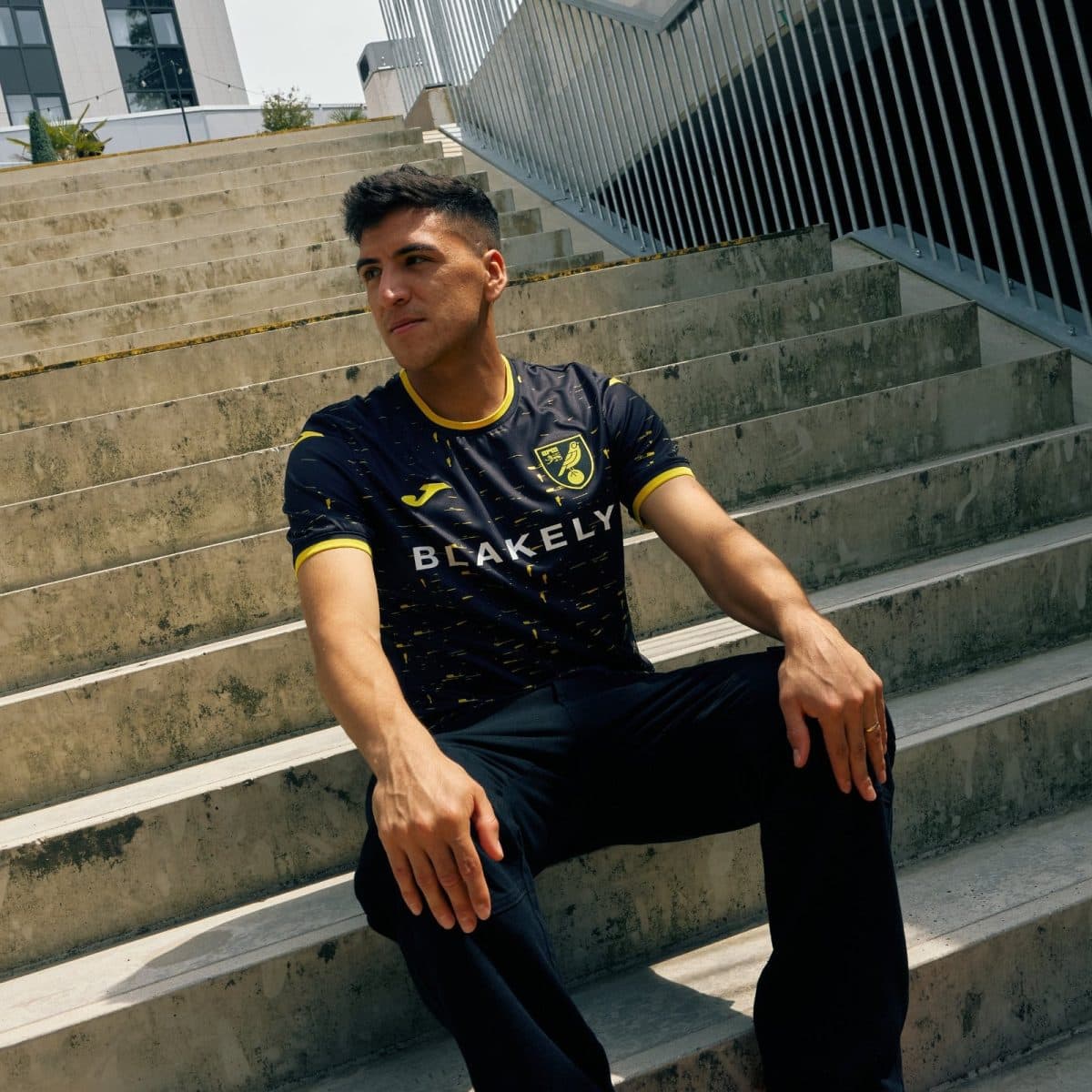 Young man sits on outdoor concrete steps in a modern urban area, wearing a black and yellow athletic jersey that says 'BLAKELY'.