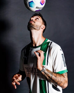 Man in white and green soccer jersey balances a ball above his head, looking up at it in a studio.