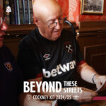 Man in a black Betway shirt signs papers, with the 'Beyond These Streets' Cockney Kit 2024/25 logo visible.