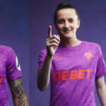 Smiling woman in a purple team jersey points toward the viewer, against a purple background.