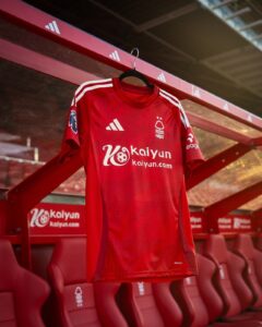 Red football jersey with white stripes on a hanger in a dugout, featuring Kaiyun sponsor and a forest club crest on the chest.