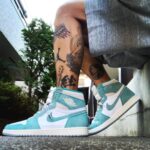 Teal and white high-top sneakers on a tattooed leg, outdoors on a concrete surface.