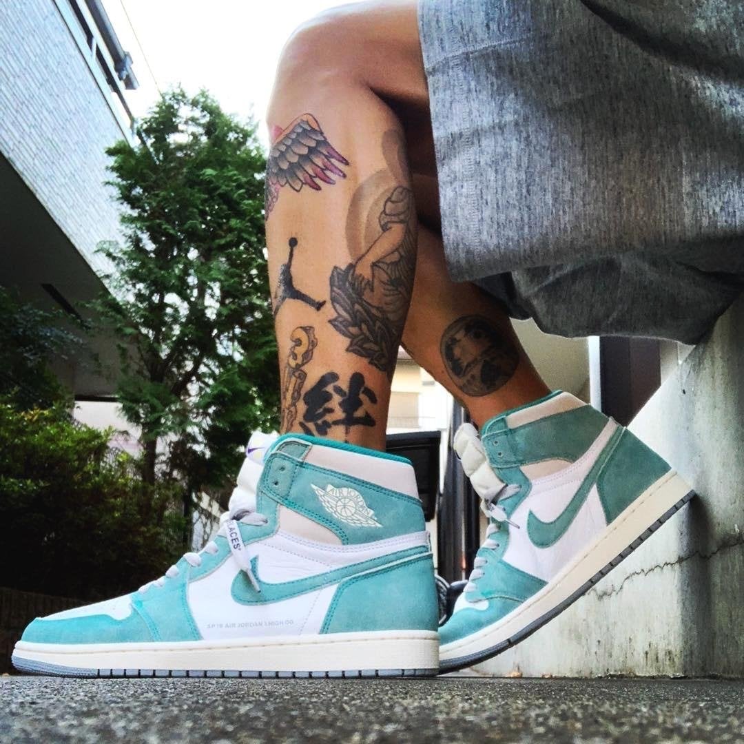 image_0eeb098e-af99-4593-9b56-3c8b78f69af7 Teal and white high-top sneakers on a tattooed leg, outdoors on a concrete surface.