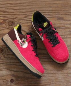 Bright pink Nike sneakers with white swoosh, burgundy accents, black-and-white laces, and a yellow heel pull tab on a wooden floor; a small Minnie Mouse charm hangs from one lace.