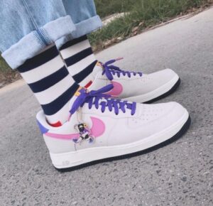 White sneakers with purple laces and pink swoosh, decorative charm, worn with black-and-white striped socks on a sidewalk.