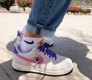 Close-up of white Nike sneakers with purple laces and pink side stripe, worn with cuffed jeans and white socks; a small cartoon charm hangs from the laces.