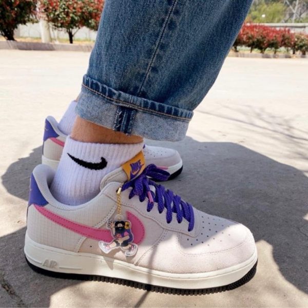 Close-up of white Nike sneakers with purple laces and pink side stripe, worn with cuffed jeans and white socks; a small cartoon charm hangs from the laces.