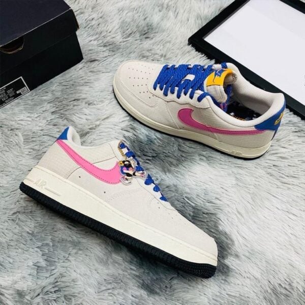 White sneakers with pink Nike swoosh, blue laces, and Minnie Mouse charms on a fluffy gray rug, with a black box and tablet nearby.