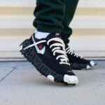 Close-up of black Nike sneakers with white laces and a speckled chunky sole, worn by a person in green pants on a concrete surface near stairs.