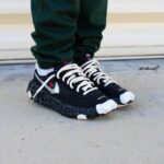 Close-up of black sneakers with chunky spiked soles and white laces, worn with green pants, standing on a concrete surface.