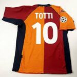 Red and orange striped football jersey with the name 'TOTTI' and number '10' on the back, featuring a UEFA Champions League patch on the right sleeve