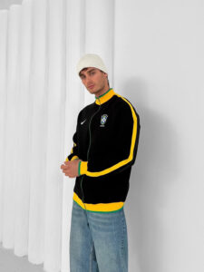 Young man modeling a Brazil track jacket: black with yellow/green stripes, white beanie, light jeans, posing against a white wall.