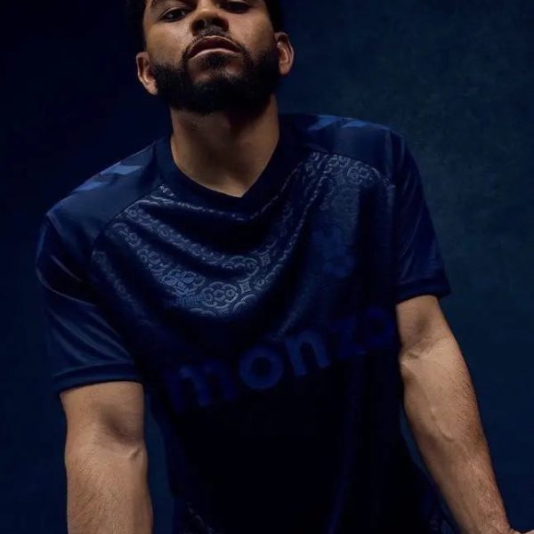 Portrait of a bearded man in a dark blue patterned shirt, leaning toward the camera against a moody blue background.