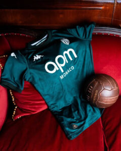 Teal AS Monaco football jersey laid on a red velvet couch with a brown vintage soccer ball beside it.