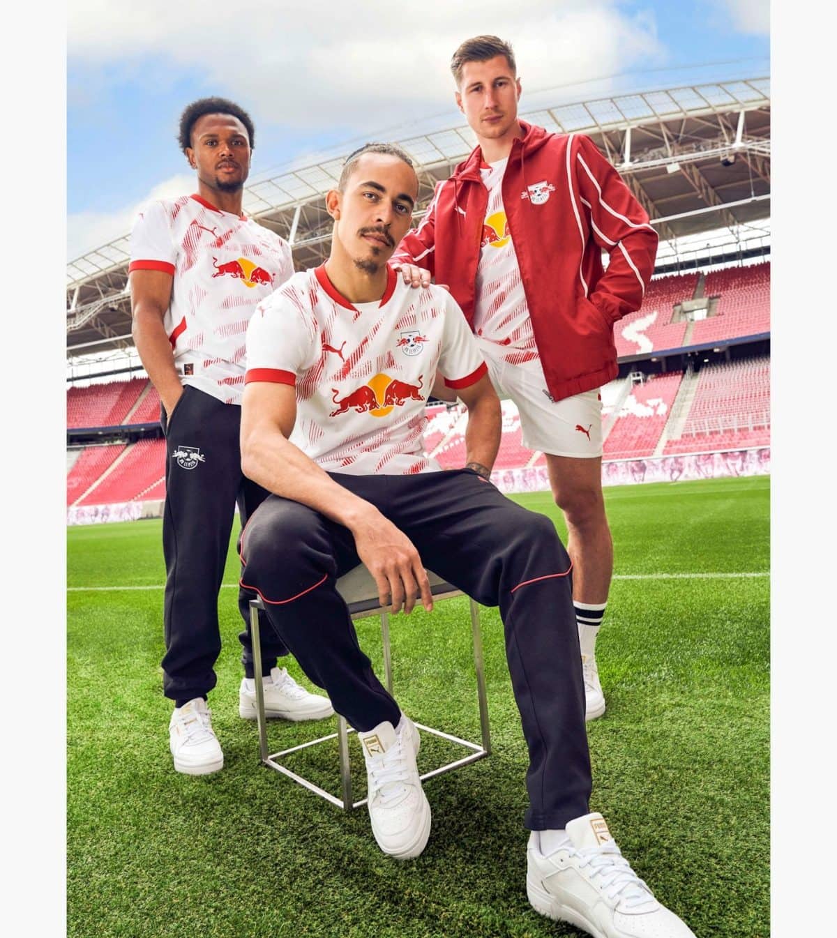 Three athletes pose on a soccer field wearing Red Bull gear in a stadium. A man sits on a metal stool while two stand beside him, all in white and red uniforms.