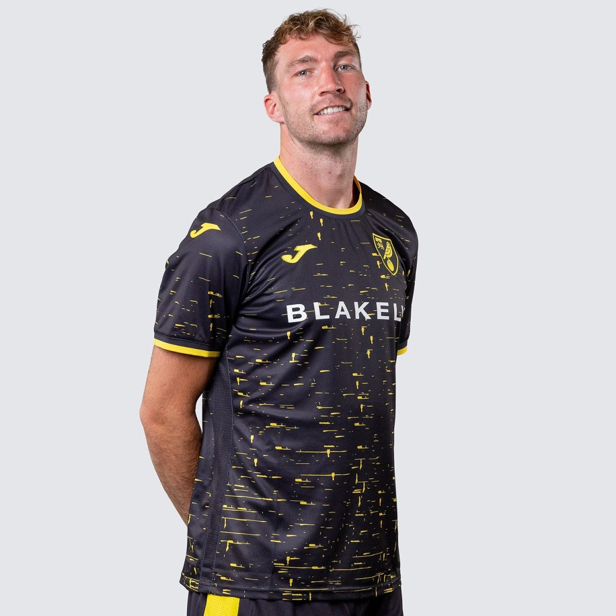 Man wearing a dark navy football jersey with yellow speckle pattern, yellow trim, crest on the chest, and 'BLAKELY' across the front against a light gray background.