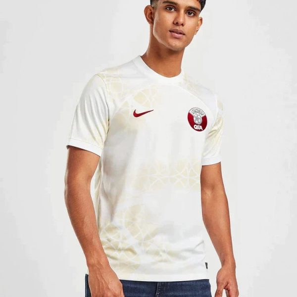 Young man wearing a white soccer jersey with a red Nike swoosh on the left chest and a circular crest on the right chest, standing against a light background.
