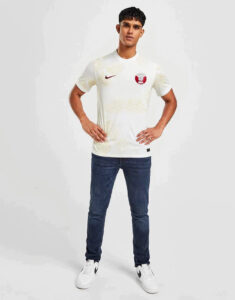 Young man modeling a white athletic jersey with a red circular logo on the left chest, paired with dark jeans and white sneakers.