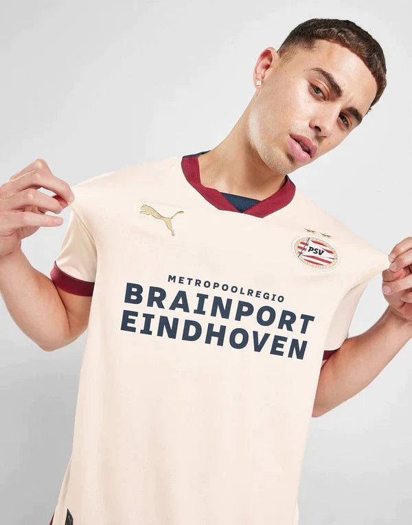 jd_650587_a_e44747ba-a5dc-4431-ae5a-353b61a2f7fd | UAE SPORT STORE Male model wearing a cream PSV Eindhoven football jersey with maroon trim, displaying the team slogan Brainport Eindhoven and the PSV badge.