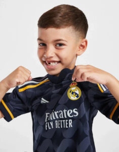 Smiling young boy posing with a Real Madrid jersey, showing a fist gesture as if ready to play.