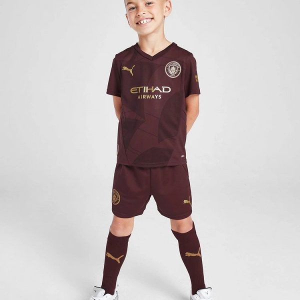 Young boy in a maroon soccer kit, standing with hands behind back, smiling at the camera.
