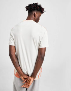 Back view of a man in a cream T-shirt with orange piping and gray shorts, hands clasped behind his back.