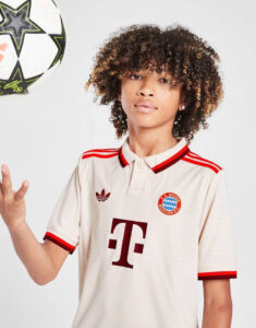 Young person with curly hair wearing a cream FC Bayern Munich jersey with red trim, touching a soccer ball in midair to the left.