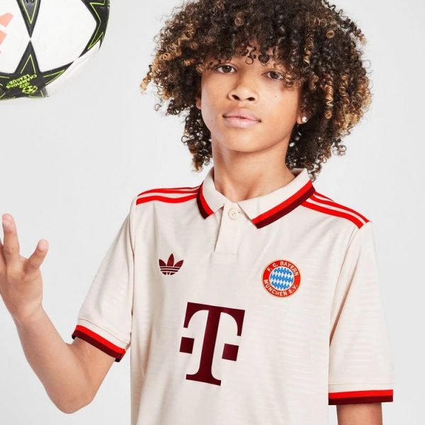 Young person with curly hair wearing a cream FC Bayern Munich jersey with red trim, touching a soccer ball in midair to the left.