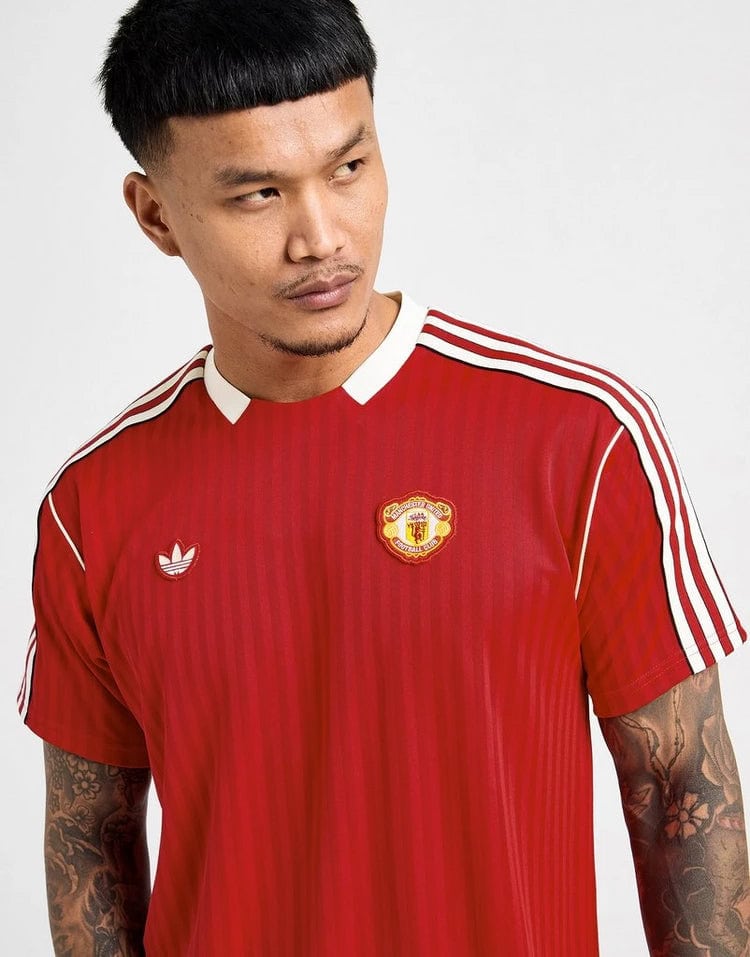 jd_749355_b | UAE SPORT STORE Man wearing a red Manchester United football jersey with white stripes on the sleeves, looking to the right.