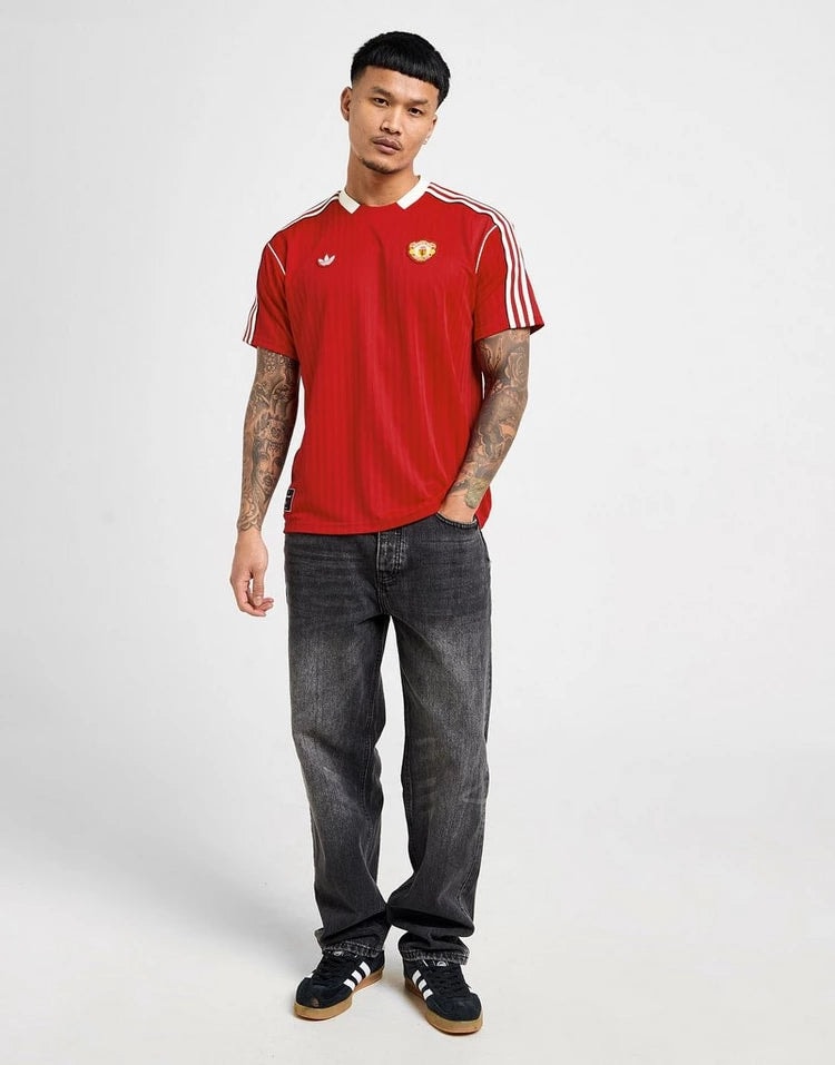 jd_749355_d | UAE SPORT STORE Man wearing a red Manchester United jersey with white stripes, dark jeans, and black sneakers, standing against a light background.