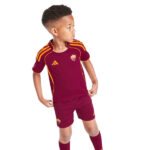 Young boy wearing maroon AS Roma soccer kit with orange accents, standing with hands on hips and looking to the right.