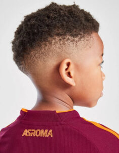 Boy shown from the back wearing a maroon Roma jersey with an orange trim; short curly fade hairstyle visible.