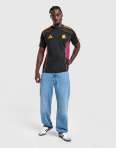 Man posing in a black athletic t-shirt with yellow piping, pink side panels, blue jeans, and white sneakers against a light background.