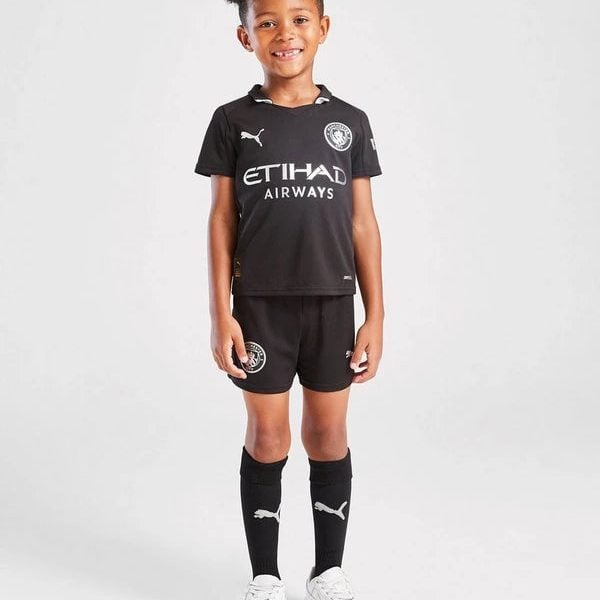 Young boy in a black soccer kit with Etihad Airways sponsor, standing and smiling on a white background, white sneakers visible.