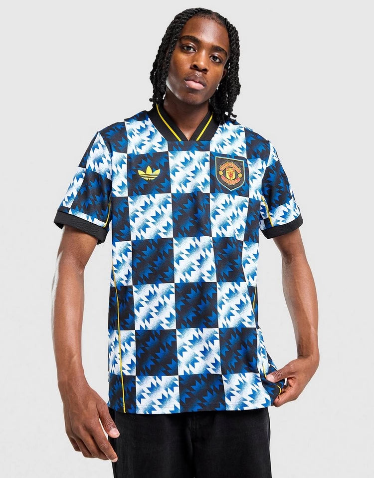 jd_760195_b Person wearing a blue and white geometric Manchester United football jersey with Adidas logo and crest, standing against a light gray backdrop.