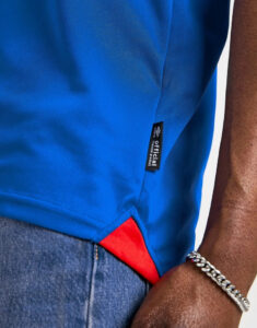 Close-up of a bright blue athletic shirt with a small black tag reading 'Official', showing a red inner hem at the bottom, worn over denim jeans and a silver chain bracelet on the wrist.
