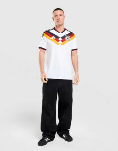 Man wearing a white Germany national football team jersey with black jeans and sneakers, standing in a neutral studio background.