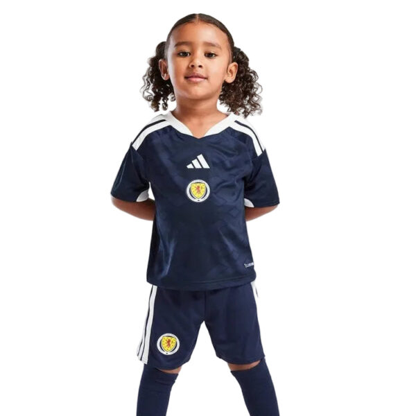 Young girl in a navy soccer uniform with white stripes and a Scotland crest, standing with hands behind her back.