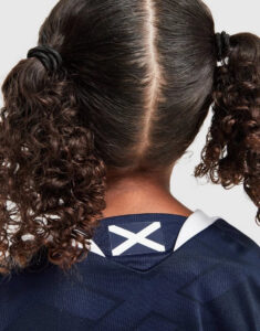 Back view of a person with curly pigtails, wearing a navy sports jersey with a Scottish flag patch at the collar edge.