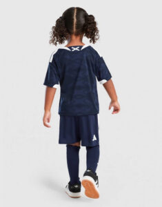 Back view of a child wearing a navy soccer jersey and shorts, navy socks, and black Adidas sneakers on a light background.